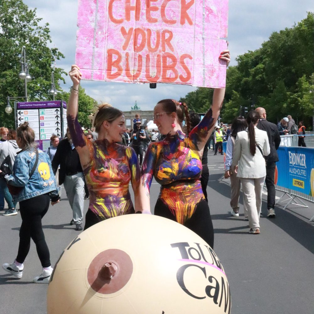 Foto vom VitaminWell-Frauenlauf. Zwei Buusenkollektiv-Mitglieder halten ein Schild mit „Check your buubs" hoch.